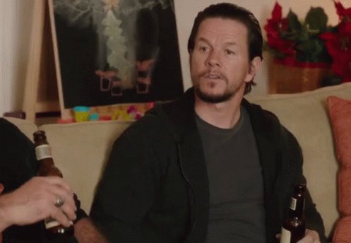 The image features a man sitting on a couch with two beer bottles, one of which has a label that reads "Nakamigos". The man appears to be holding the beers while looking at something off-camera or engaged in conversation. The background includes a potted plant and an art piece hanging on the wall above it.