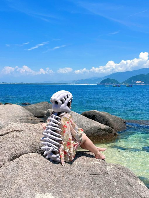 A person wearing a skeleton costume with a skull headpiece and floral-patterned kimono sits on rocks by the sea, looking out at the blue water and mountains in the distance.