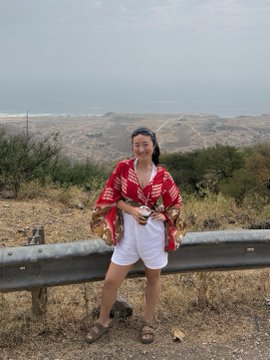 A woman sits on a roadside guardrail overlooking a scenic ocean view, smiling while wearing a red patterned top and white shorts. The uplifting coastal scene complements the post's message about finding renewed purpose and motivation through supportive community messages, contrasting the negative feelings about the crypto industry with a moment of personal reflection and gratitude.