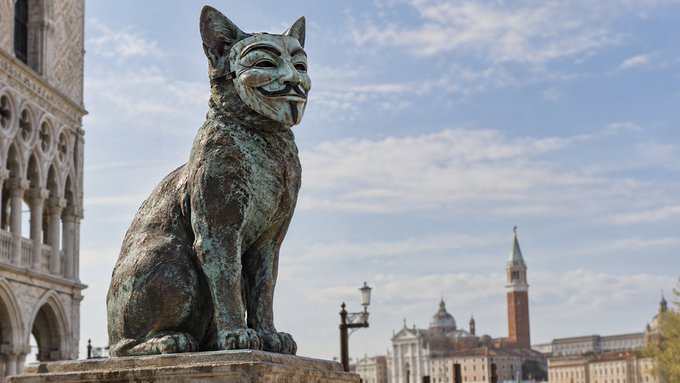 A bronze statue of a smiling, masked cat sits in an Italian plaza with historic architecture and a tower visible in the background under a partly cloudy sky.