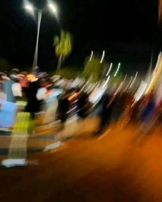 A blurry nighttime photo of a crowd gathered in a street or plaza, with people standing around under streetlights. The image's chaotic, motion-blurred scene pairs with the post's joke about watching a crypto/financial portfolio drop to nearly nothing, capturing the frantic energy of witnessing one's investments tank.