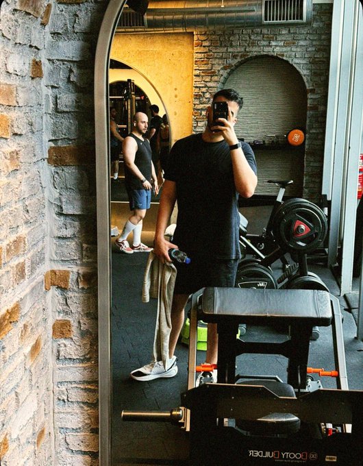 A man takes a mirror selfie with his phone in a gym, holding a water bottle and towel, with gym equipment and another person visible in the reflection.