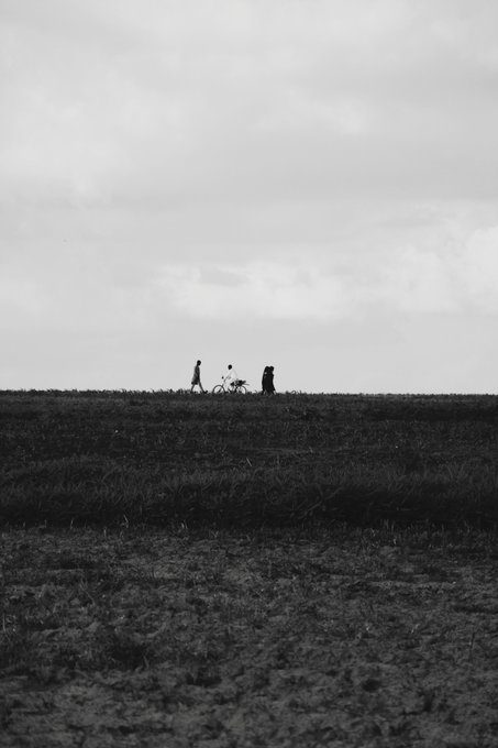 A black-and-white photograph showing silhouettes of two people with a bicycle walking along the ridge of a field under a vast gray sky.