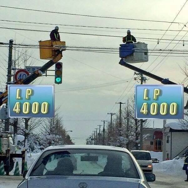 Two workers in cherry pickers service overhead power lines above a snowy street, while digital signs display "LP 4000" resembling a stock ticker. A white car drives beneath them past a green traffic light.