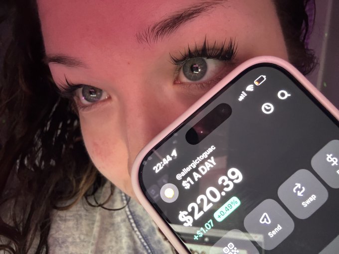 A close-up selfie shows a person holding a smartphone displaying a meme token chart for $1ADAY with a value of $220.39, up +$1.07 (+0.49%).