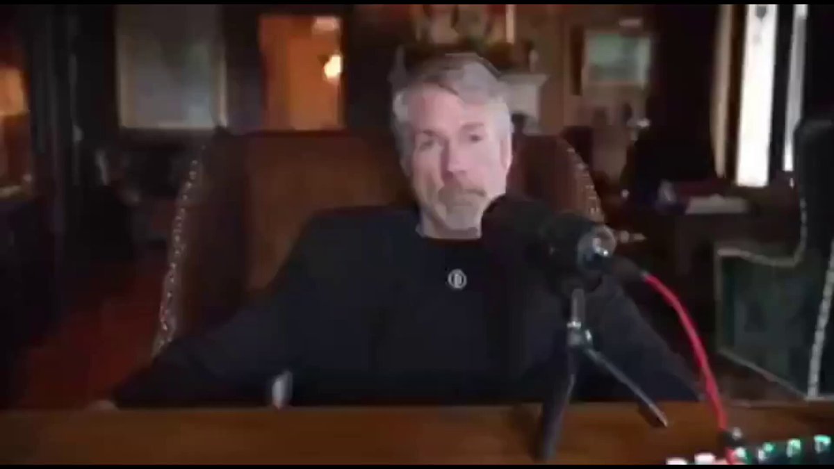 The image shows a man sitting at a desk with a microphone in front of him, suggesting that he might be hosting or participating in an interview or podcast recording session. The man appears to be wearing a black shirt, which contrasts with his white hair. He is seated on a chair and positioned behind the desk, giving viewers a clear view of him as the main subject of the image.