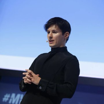 A man in a black shirt giving a presentation on stage, wearing a small headset microphone with a blue background behind him.