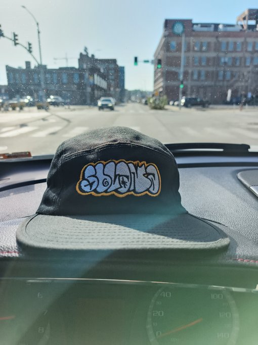A gray graffiti-style snapback hat rests on a car dashboard, with a city street and brick buildings visible through the windshield.