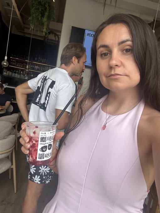 A woman in a pink tank top takes a selfie at a bar while holding a plastic cup containing a berry-colored drink.