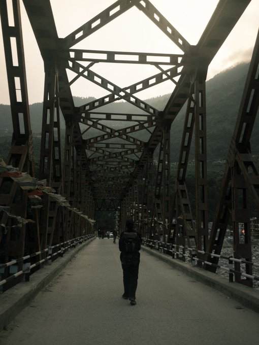 A person with a backpack walks along a steel truss bridge, with prayer flags hanging from the side and mountains visible in the background.