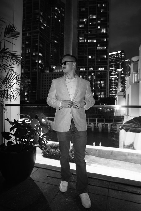 A black-and-white photo shows a man in sunglasses and a light blazer standing on a balcony at night, adjusting his jacket button with a city skyline of illuminated high-rise buildings in the background.