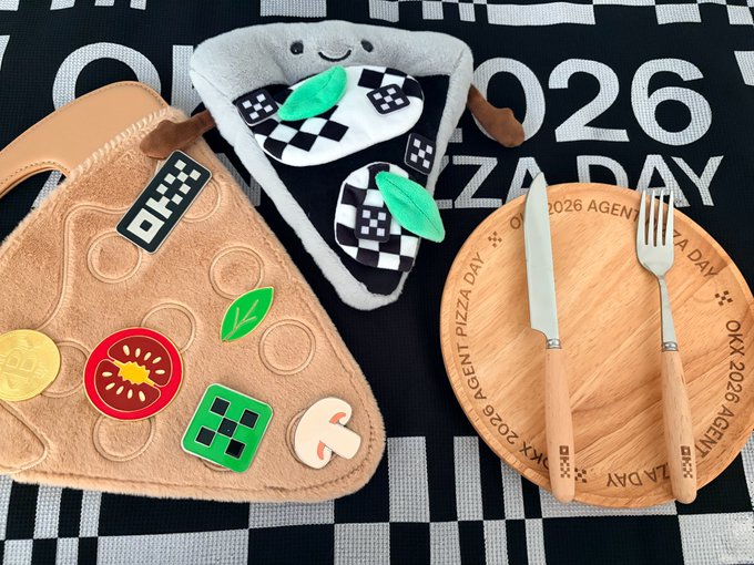 The image features a black and white checkered tablecloth with various items placed on top of it, including a pizza slice, a plate, a fork, a knife, and some stickers. The pizza slice appears to be made out of fabric or stuffed animal material, adding a playful touch to the scene. There are also two stickers visible in the image, one located near the bottom left corner and another towards the top right corner.

The table setting includes a plate with a fork and knife on it, as well as a pizza slice placed next to them. The overall arrangement of items suggests that someone has prepared for a casual meal or gathering, possibly involving pizza as the main course.