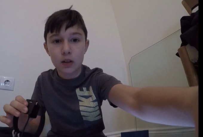 A young boy with dark hair, wearing a grey t-shirt with 'TRAINING' printed on it, sits indoors holding a black wired device, possibly a webcam or microphone.