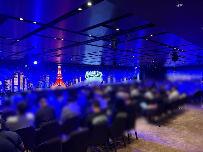 An indoor event space, likely a corporate gathering or conference, features a large screen displaying a Tokyo cityscape with Tokyo Tower illuminated, while the audience is blurred in the foreground.