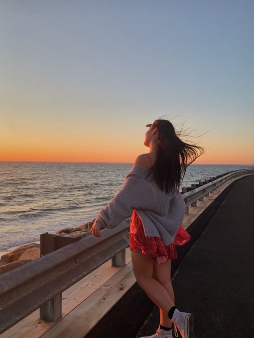 The image features a woman standing near a railing, overlooking the ocean at sunset. She appears to be enjoying her time by the water, possibly taking a break from work or simply appreciating the beautiful view. The photo captures the essence of the moment with its warm colors and serene atmosphere.