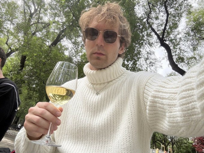 A selfie of a man wearing sunglasses and a cream-colored turtleneck sweater, holding a glass of white wine outdoors with trees in the background.