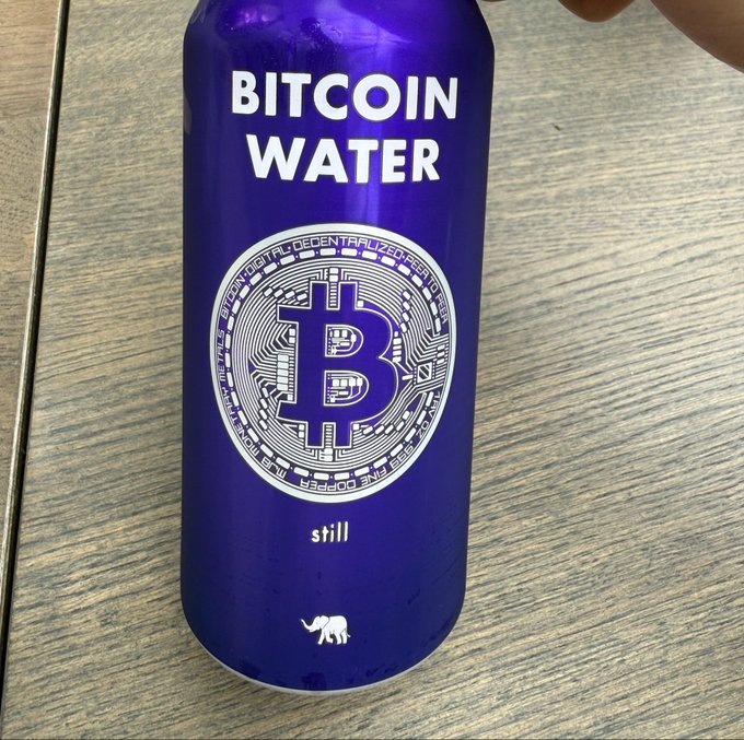 A purple bottle of 'Bitcoin Water' still water featuring a large Bitcoin 'B' logo with circuit board details and an elephant icon at the bottom.