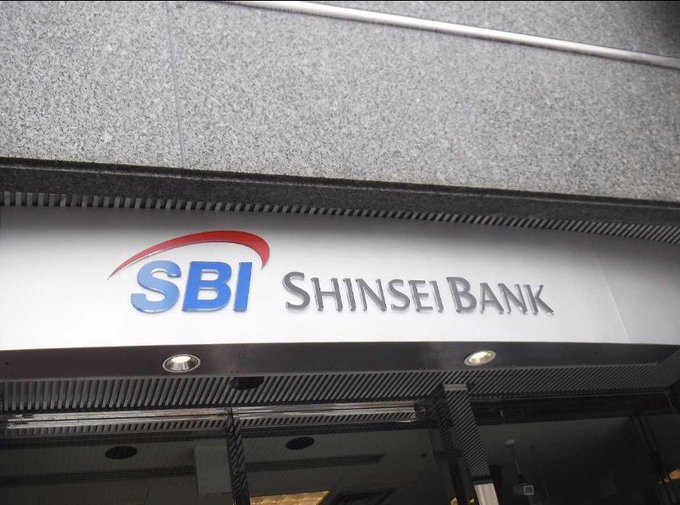 The image shows a building with a large sign that reads "SBI Shinsei Bank" above its entrance, indicating the bank's name or logo. The sign is prominently displayed for all to see. Below the sign, there are two lights on the side of the building, which could be used for security purposes or as an advertisement.