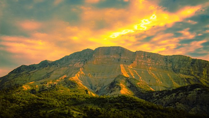 A scenic photograph of a mountain landscape at sunset, with golden-orange clouds filling the sky above rugged peaks.