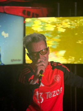 The image features a man wearing an orange shirt, standing in front of two large screens displaying images or videos. The man appears to be singing into a microphone, possibly performing at a party or event. The background includes a TV screen and another smaller screen, which could also be part of the entertainment setup for the gathering.