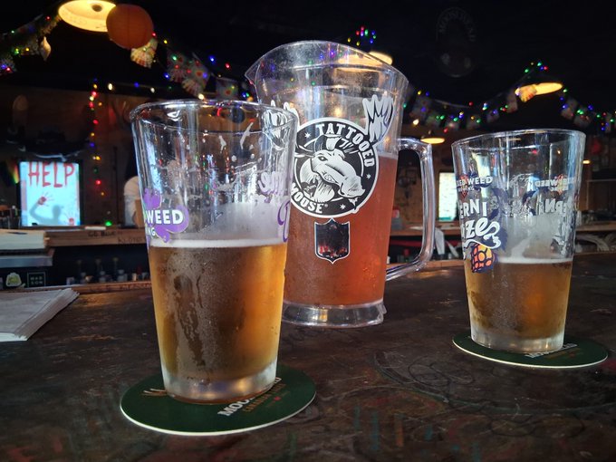Two pint glasses of beer and a larger mug of amber-colored beverage sit on coasters on a wooden bar table; the glasses are decorated with stickers and graffiti, set against a dimly lit bar background with string lights.