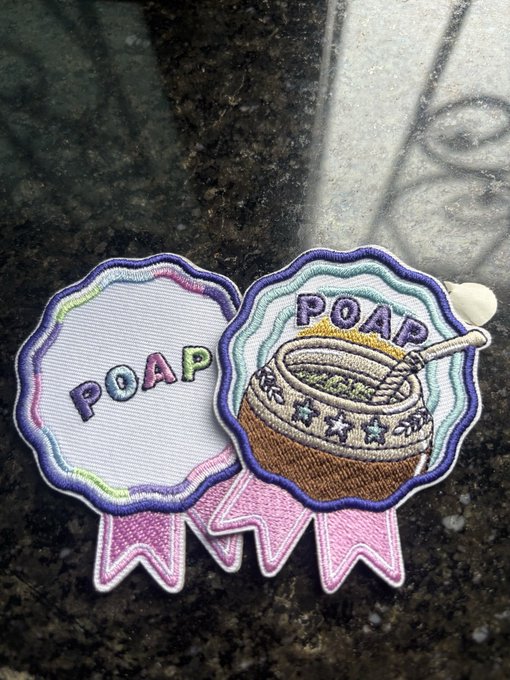 Two POAP (Proof of Attendance Protocol) embroidered patches rest on a dark surface, each featuring the text "POAP" and shaped like a ribbon medal—one displaying a bowl of food with a straw and the other being a blank version, both fitting the user's mention of hoping for an Indian-themed POAP for the November event.