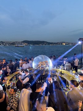 The image shows a large group of people gathered at an outdoor event, possibly a party or festival. The crowd consists of men and women standing close together, with some individuals holding drinks in their hands. There are also several bottles scattered throughout the scene, indicating that beverages have been consumed by attendees. In addition to the people and drinks, there is a large glass ball present at the center of the gathering, which could be part of an entertainment or decoration for the event. The overall atmosphere appears lively and social, with everyone enjoying themselves in the outdoor setting.