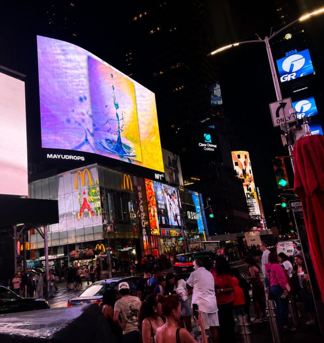 A nighttime street scene in Times Square featuring a large billboard displaying a yellow artwork labeled 'MAYUDROPS' above a McDonald's, with 'NFT' visible nearby.