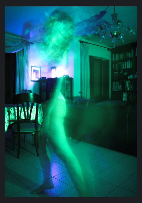 An atmospheric, blurry long-exposure photograph shows a ghostly green figure moving through a dimly lit living room with a chandelier and dark furniture.