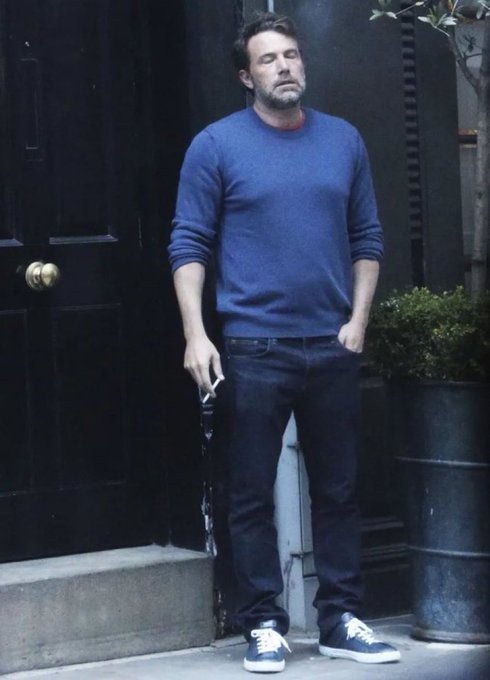 A man with a beard wearing a blue sweater, dark jeans, and blue sneakers stands with eyes closed on a sidewalk next to a black door, with a potted plant in a metal barrel to his right.