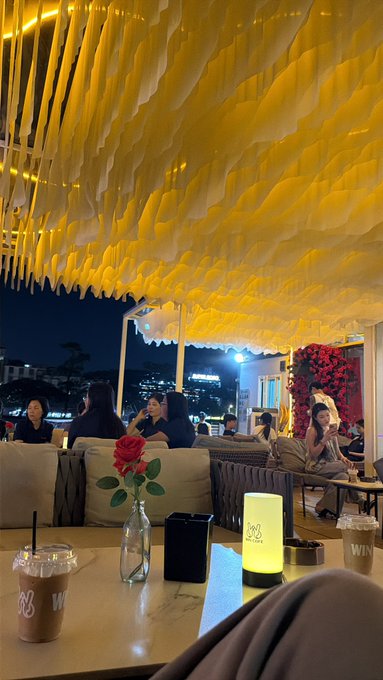 The image shows a group of people sitting at tables inside an outdoor restaurant, enjoying their time together under a large yellow canopy. There are several chairs around dining tables with cups and vases placed on them. A potted plant can be seen near one of the tables, adding some greenery to the scene. The photo captures the atmosphere of a social gathering or event taking place in an outdoor setting during nighttime hours.