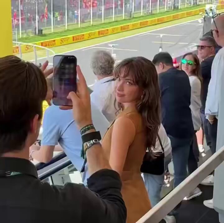 In the image, there are multiple people at an event taking photos using their phones. One man, wearing a black shirt, is seen capturing a picture of someone standing next to him with his cell phone. Another person nearby also has their cell phone in hand, possibly taking photos as well. The crowd appears to be engaged and interested in the ongoing photo session. In the background, there are race tracks on which the attendees seem to gather for the event. The presence of numerous people and the focus on capturing memories during this gathering indicate that the image is related to an event or race track where cell phones have become a common tool for sharing experiences with others.