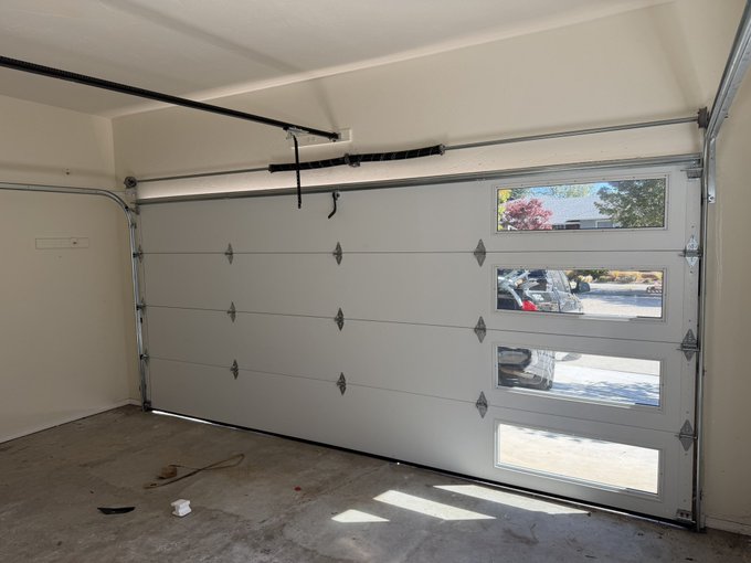 A clean, empty garage interior featuring a modern white sectional garage door with multiple windows showing a view of a driveway and neighboring houses outside.