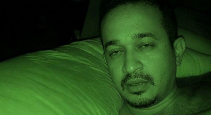 A close-up selfie of a man with short hair and a goatee, resting his head on a pillow with a green night-vision filter applied to the image.
