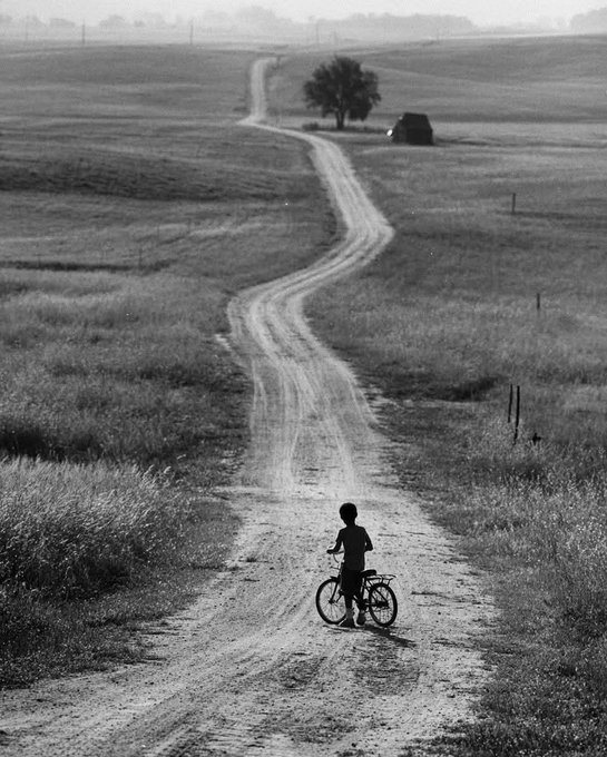 A black-and-white photograph shows a young person riding a bicycle down a long, winding dirt road through open countryside toward a distant tree.