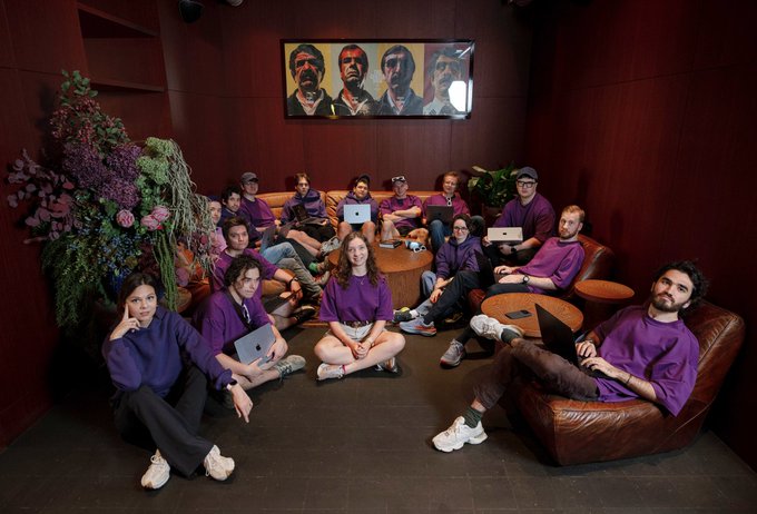 A group of around fifteen people in matching purple tops sit and lounge on brown leather sofas in a wood-paneled room, many working on laptops, with a four-panel portrait painting of bearded men hanging on the wall behind them and a large floral arrangement to the left—evoking a cohesive startup or AI team collaborating together, fitting the post's theme of multiple teams/models working as one unified system.