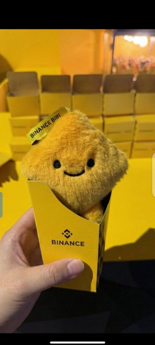 A hand holding a yellow Binance-branded box containing a plush 'Binance BIBI' mascot, with a stack of similar yellow boxes in the background.