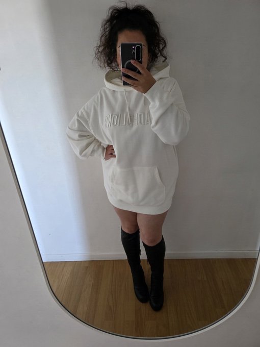 A woman with curly hair takes a mirror selfie while wearing an oversized white 'Balenciaga' hoodie and black knee-high boots.