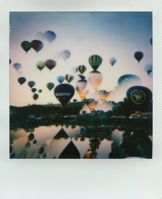 A Polaroid-style photo shows numerous hot air balloons of various designs floating above a landscape at dawn or dusk.