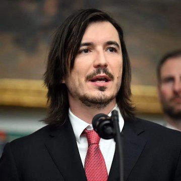 A man with long dark hair, a goatee, and a red tie speaks at a microphone, appearing to give a speech or address an audience.