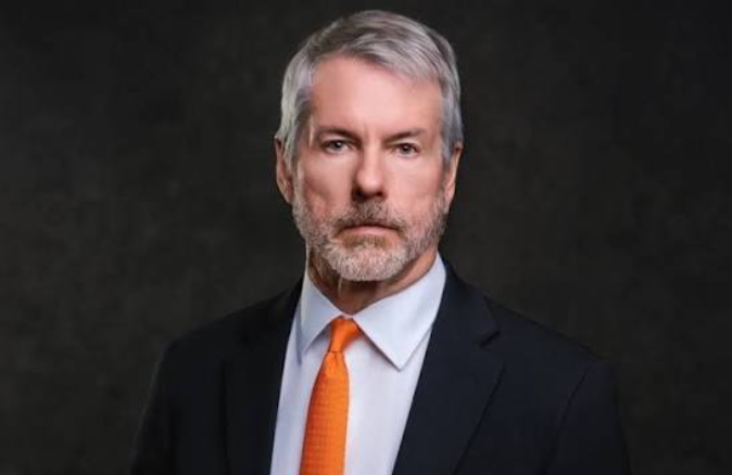 A distinguished man with gray hair and a beard, wearing a dark suit and bright orange tie, poses for a formal portrait against a dark background. The image likely depicts Michael Saylor, whose recent large Bitcoin (BTC) transfer of $297M has sparked speculation in the crypto community about potential market moves.