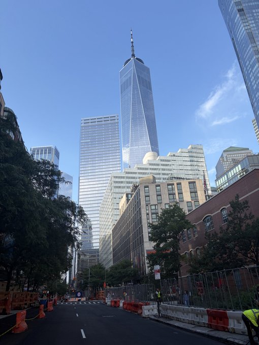 A street-level view looking up at the One World Trade Center and surrounding skyscrapers in New York City on a clear day, with road construction visible in the foreground.