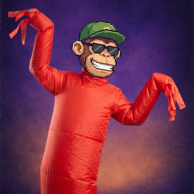 A person wearing a bright red full-body suit and a pixelated mask resembling a retro video game character with sunglasses, a green cap, and a big smile poses with their hands raised like claws against a purple and orange background.