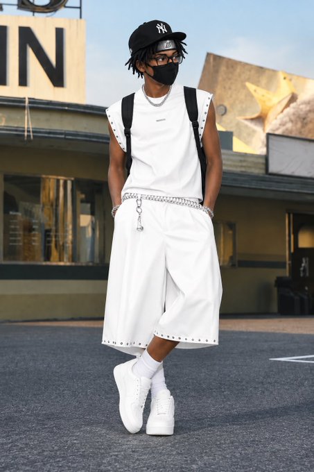 A young person modeling streetwear, wearing a black NY Yankees cap, black face mask, white sleeveless hoodie, white shorts with stud details, a chain belt, and white sneakers, standing outdoors near a storefront.