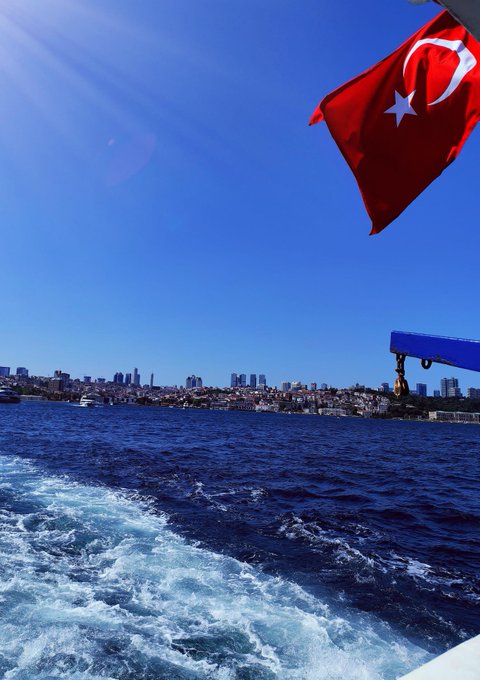 A Turkish flag waves proudly from the deck of a ferry as the boat cuts through the Bosphorus waters, with Istanbul's skyline visible in the distance under a clear blue sky—evoking the resilience and quiet triumph of those who held onto belief through turbulent times.