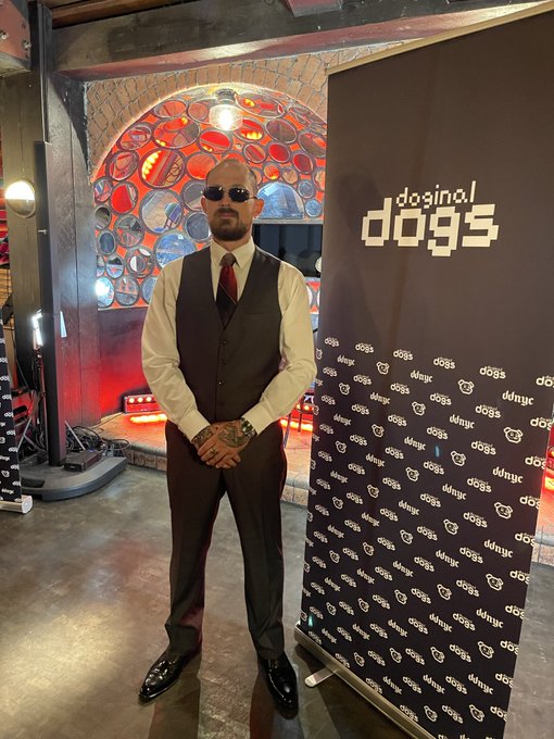 A bald man wearing sunglasses, a vest, white shirt, and red tie poses at a 'doginal dogs dognyc' branded event backdrop with stained glass windows visible behind him.