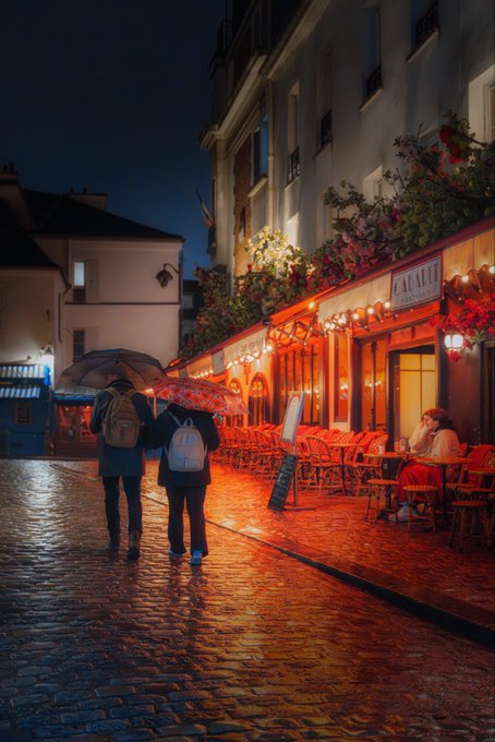 An atmospheric nighttime photograph of a couple walking under umbrellas down a wet cobblestone street past a warmly lit restaurant named 'CAFAUX'.