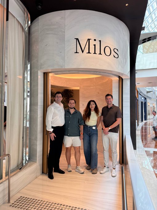 Four people pose for a photo in front of the entrance to the 'Milos' restaurant, with the name displayed in large black letters on a marble wall.
