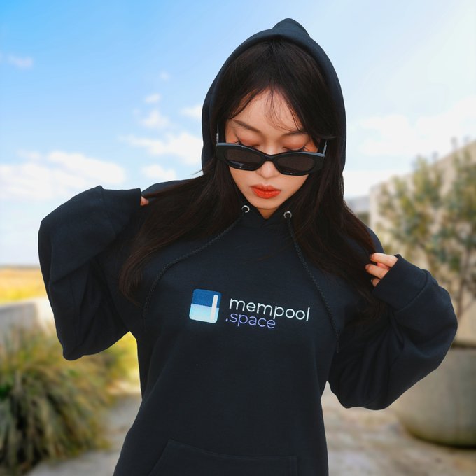 A young woman wears sunglasses and a black hoodie featuring a 'mempool.space' logo with a blue Bitcoin symbol, standing outdoors under a clear sky.