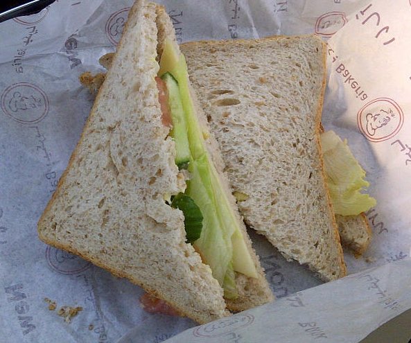 The image shows a sandwich cut into two pieces, with one piece being larger than the other. The sandwich appears to be made of bread and filled with lettuce and tomato slices. The background features a white paper wrapper that has red writing on it. The text "AVAX" is also present in the image.